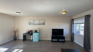 Una sala de estar con televisor de pantalla plana, una pequeña kitchenette, una mesa de comedor y un ventanal con cortinas.