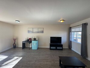 A living room with a flat-screen TV, a small kitchenette, a dining table, and a large window with curtains.