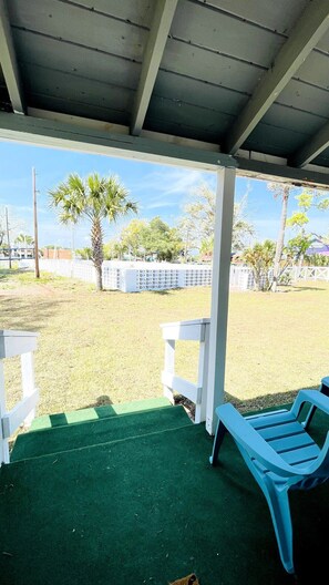 A covered outdoor area with a green artificial turf, white benches, and a view of a grassy field with palm trees.