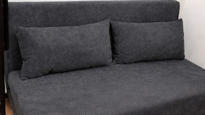 A dark grey sofa with two cushions in a corner of a room with wooden flooring and a white wall.