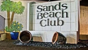 A sign for Sands Beach Club with a brick floor and potted plants.