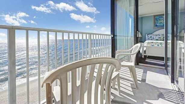 A balcony with a clear view of the sea, a bed with a blue and white bedspread, and a white chair.