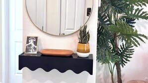 Une salle de bain avec un miroir rond, une étagère noire sur laquelle sont posés un cadre photo et un bol, une plante en pot et un tabouret en bois.