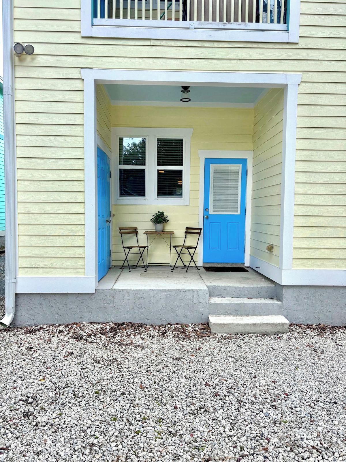Un bâtiment à deux étages, doté d’un porche à l’avant, d’une porte bleue et d’une petite table avec des chaises.