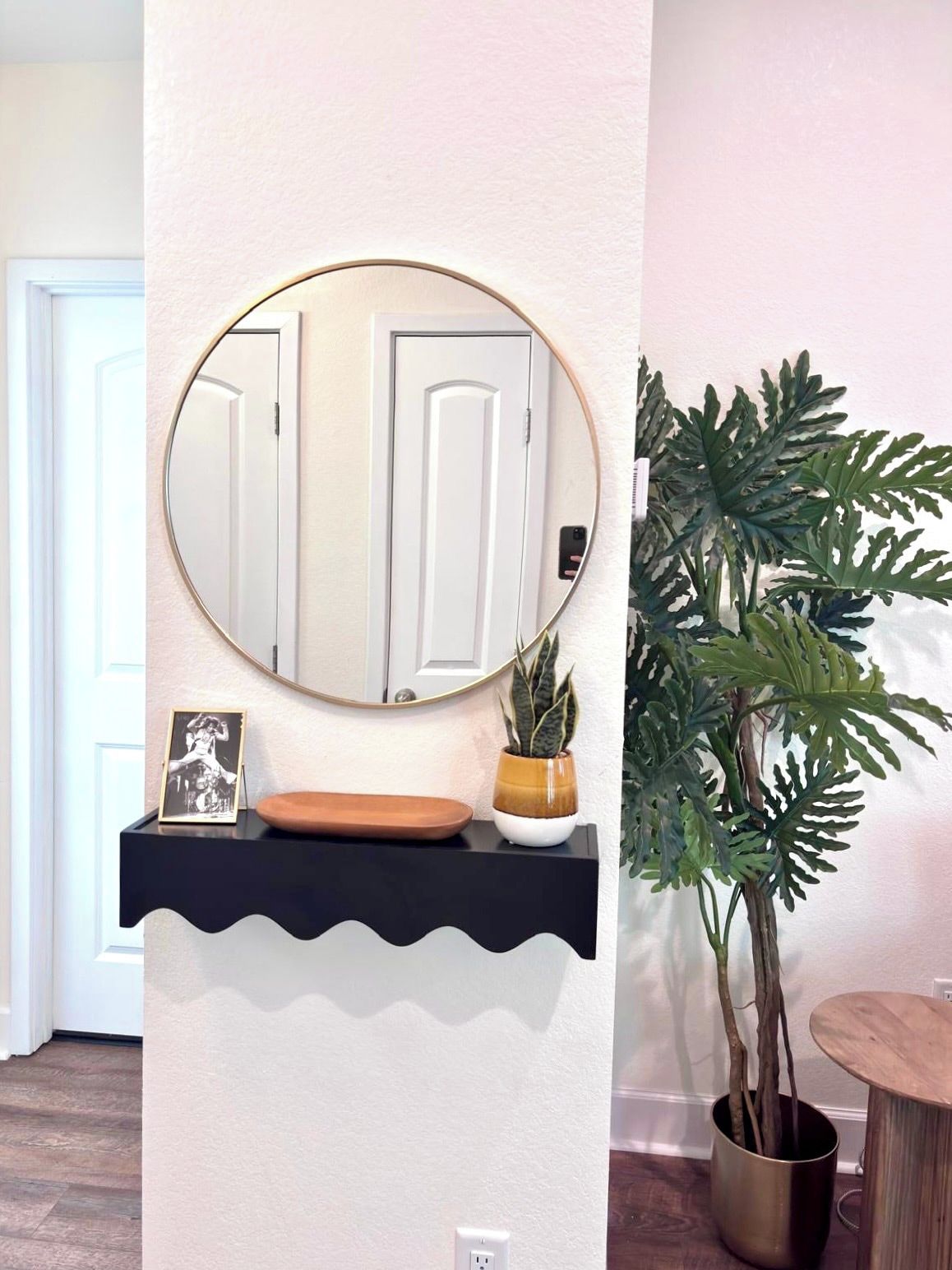 Un miroir fixé au mur, au-dessus d’une étagère noire sur laquelle se trouvent un cadre photo, une plante en pot et un petit bol en bois.