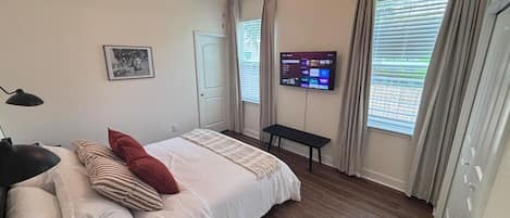 Une chambre à coucher avec un lit, un ventilateur de plafond, une télévision et une fenêtre avec des rideaux.