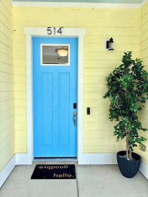 Uma porta azul com o número 514, um vaso de planta e um tapete de entrada com a inscrição 'adeus olá'.