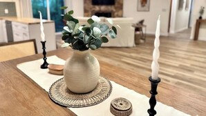 Une table à manger ornée d’un vase de feuilles vertes, de deux bougies et d’un set de table en fibres tressées, dans un salon moderne doté d’une cheminée et d’un téléviseur.