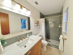 A bathroom with a double sink vanity, a toilet, a shower with a glass door, and a mirror with lights above. - Magnolia Modern, Near UF, Shands, Private (Gainesville)