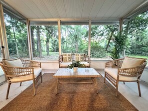 Une véranda meublée de mobilier en osier, avec une table centrale et une vue sur une zone boisée.