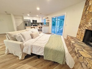 A bedroom with a bed, a stone fireplace, and a view of a kitchen and outdoor area. - Magnolia Modern, Near UF, Shands, Private (Gainesville)