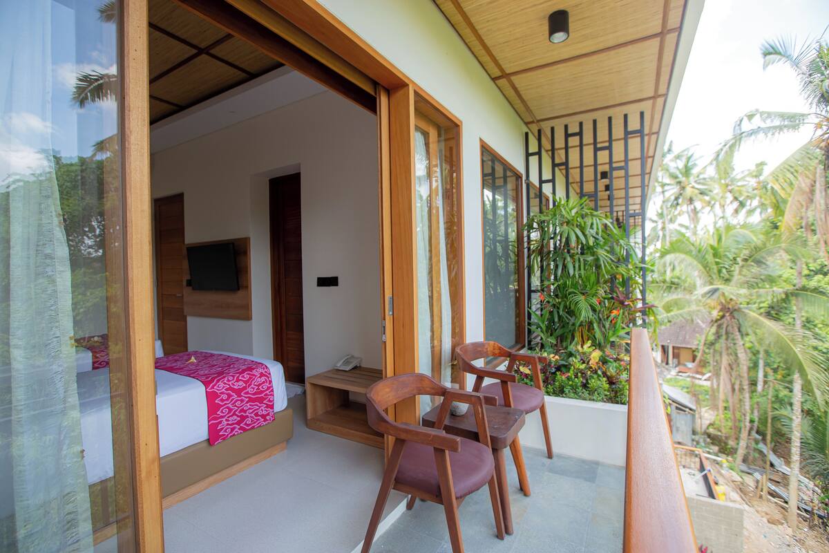 A room with a bed, a small table, and two chairs. There is a balcony with a view of palm trees and a garden.