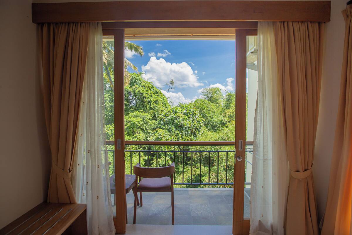 A balcony with a wooden chair and a table, overlooking a lush green landscape and a clear blue sky.