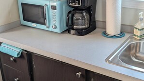 Una cocina con armarios oscuros, un microondas azul claro, una cafetera negra, un rollo de toallas de papel y una botella de jabón para manos.