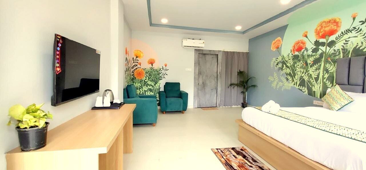 A modern hotel room with a flat-screen TV, a desk with a potted plant, a teal armchair, a bed with a large floral mural, and a window with curtains.