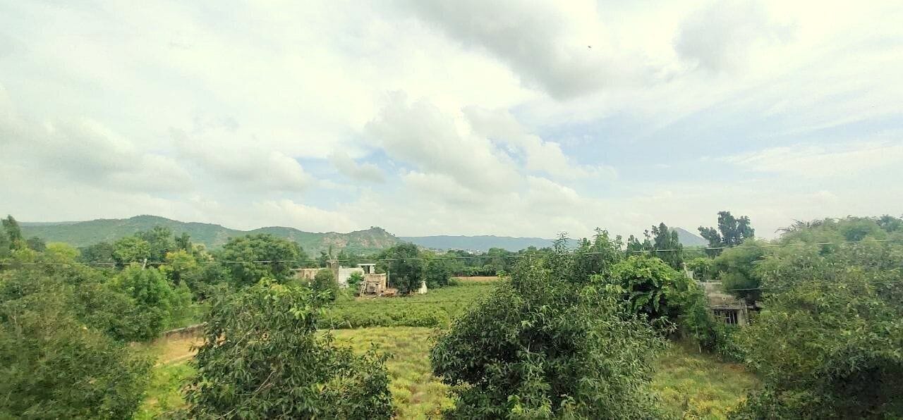 A rural landscape with scattered trees, a few buildings, and hills in the distance under a cloudy sky.