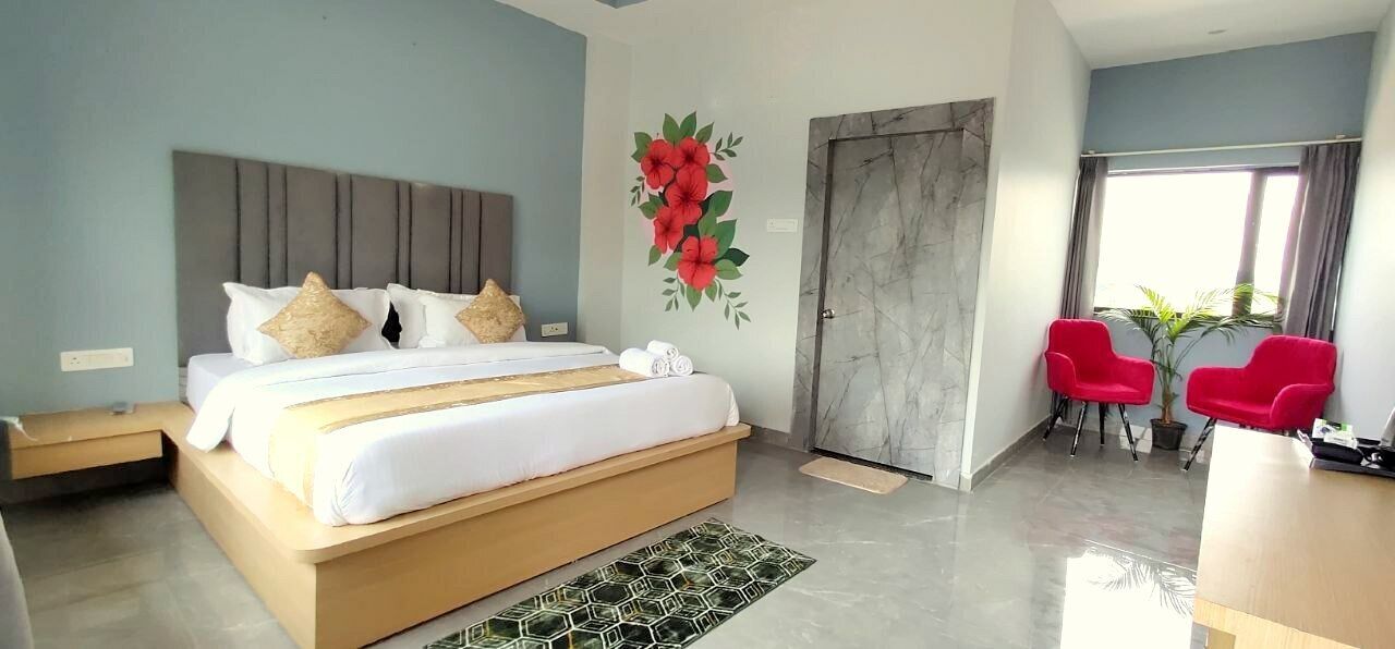 A modern hotel room with a large bed, a wooden headboard, a red floral wall decoration, a grey door, and a seating area with red chairs and a potted plant.