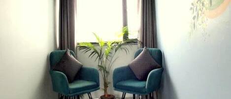 A room with a large window, two teal armchairs, a potted plant, and a wall with a leaf pattern.