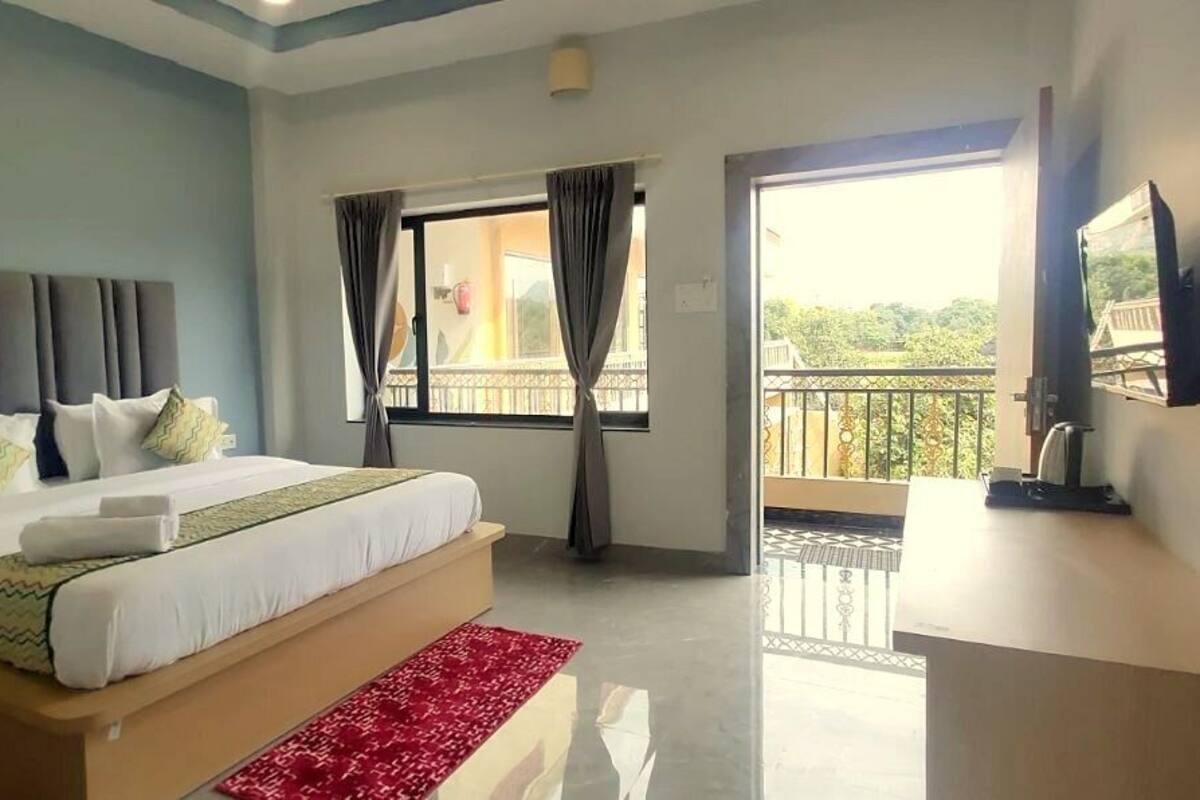 A bedroom with a large bed, a TV, a balcony with curtains, and a view of greenery.