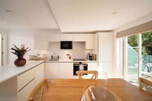 A modern kitchen with white cabinetry, a wooden dining table, and a glass door leading to a garden. - Clifftop Watch – 1-Bed River View Apartment at Tors Park (Lynmouth)