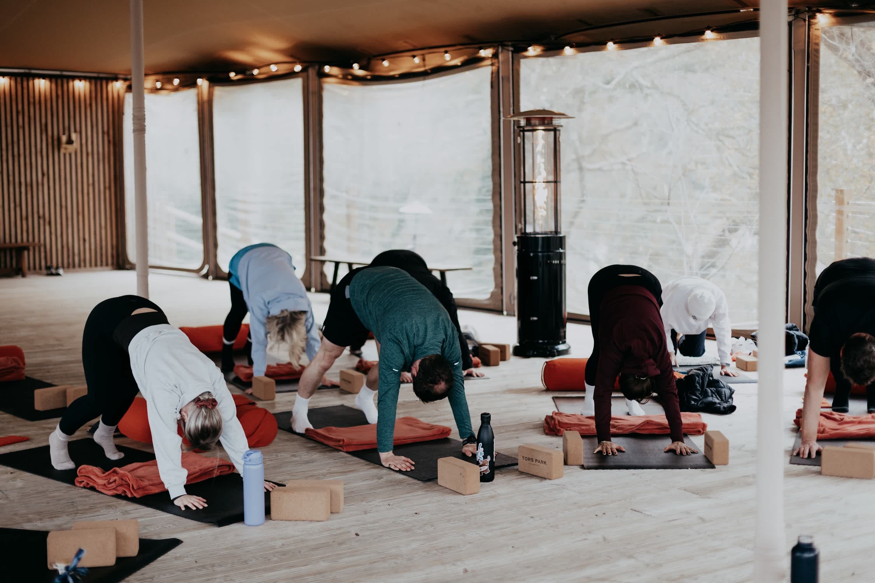 Um grupo de pessoas praticando yoga em um espaço com piso de madeira, tapetes e blocos.