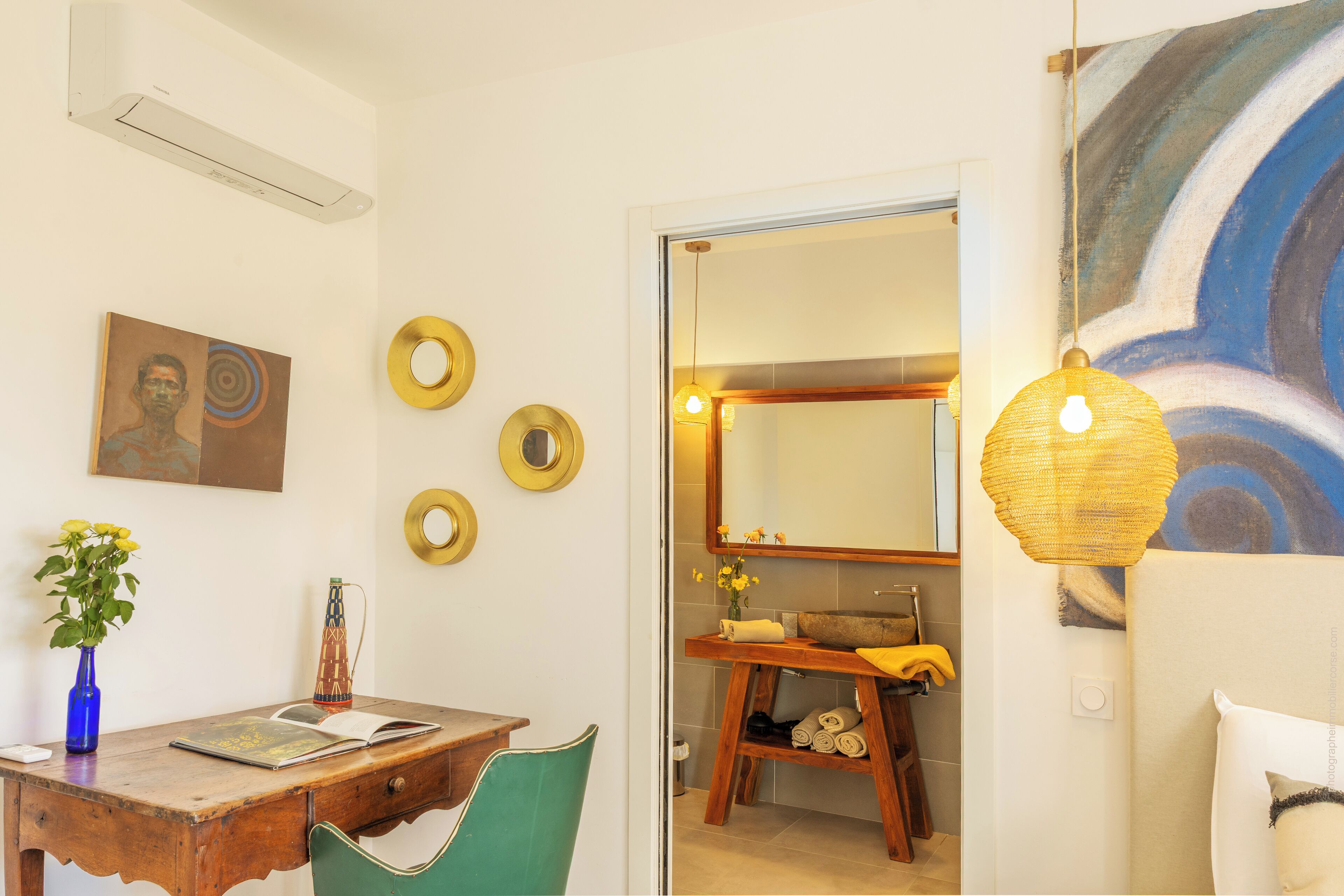 Una stanza con una scrivania in legno, una sedia verde, un vaso blu con fiori e un quadro appeso al muro.