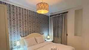 A neatly made bed with white linens, a bedside table with a lamp, patterned wallpaper, and a ceiling light fixture. - Taksim İnci Apart (İstanbul)