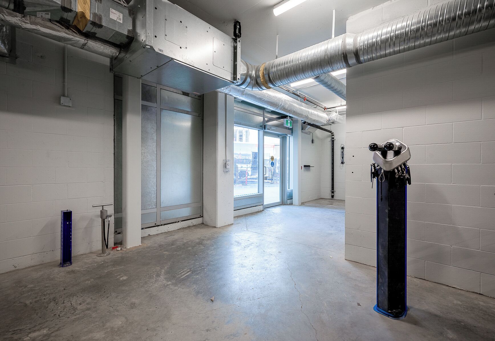 Un couloir propre, de style industriel, avec un sol en béton et des murs blancs, agrémenté d’une grande porte, d’un pilier bleu et de conduits apparents.