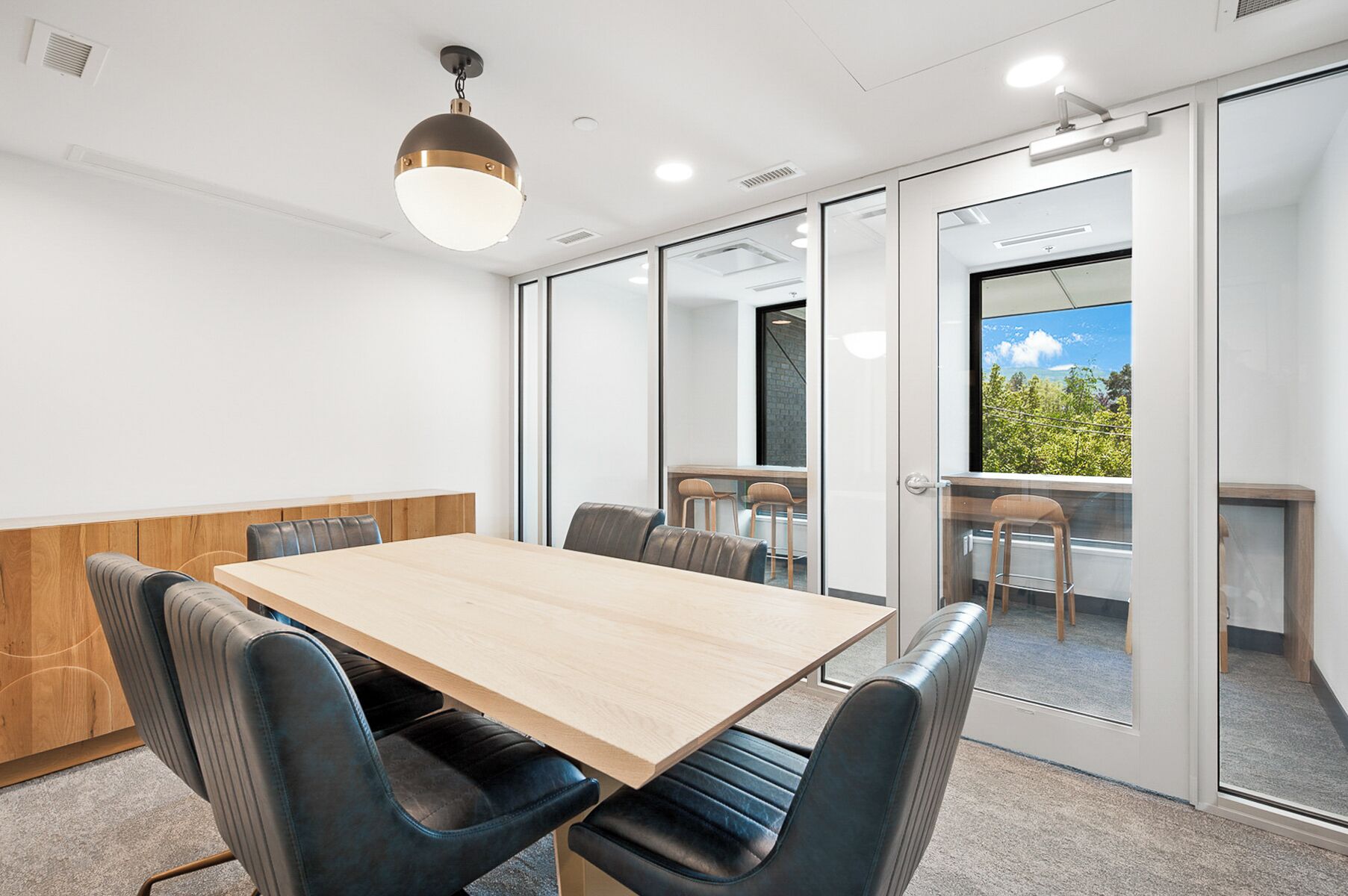 A conference room with a wooden table, black leather chairs, a hanging light fixture, and a view of greenery through a glass door.