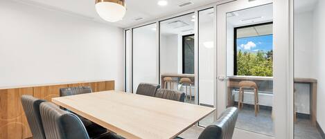 A conference room with a wooden table, black leather chairs, a hanging light fixture, and a view of greenery through a glass door.