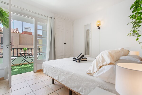 A bedroom with a bed, a bedside table, a lamp, a window, and an open door leading to a balcony with a table and chairs.