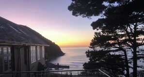 Vista costera con una casa que domina el mar, un muelle y una ladera boscosa.