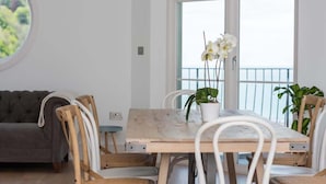 A dining area with a wooden table and white chairs, a grey sofa, and a potted plant on the table.