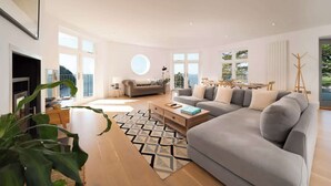 A spacious living room with a large grey sofa, a coffee table, and a TV. There are large windows providing natural light and a view of the outdoors.
