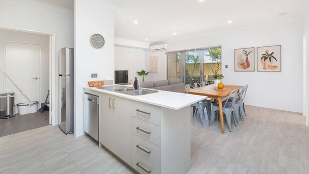 Una cocina moderna con armarios blancos, una isla central con pileta y un comedor con mesa y sillas de madera.