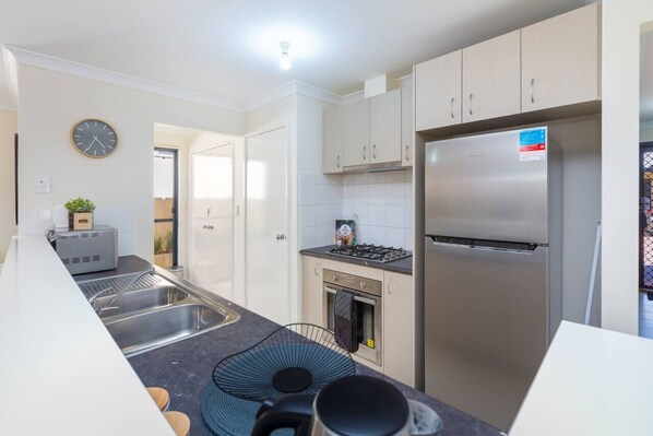 A modern kitchen with a stainless steel refrigerator, a built-in oven, and a gas stove. There is a sink with a double basin and a clock on the wall.