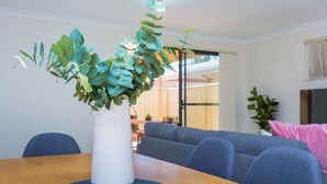 A dining table with a white vase containing green foliage, a blue placemat, and a grey sofa in the background. - Family perfection in quiet & convenient location! (Balcatta)