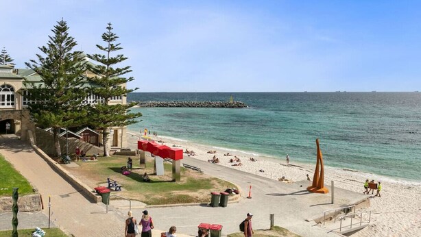 A beach with a clear view of the ocean, a red sculpture, and a building with a green roof.