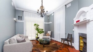 A living room with a grey sofa, a glass coffee table, a chandelier, a fireplace, and a white mantelpiece.