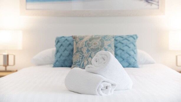 A neatly made bed with a rolled white towel, a blue patterned pillow, and a framed picture of a beach scene on the wall. - A2 - Spacious Apartment w/ Wi-Fi + Kitchenette (Connolly)