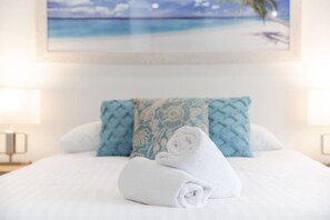 A neatly made bed with a rolled white towel, a blue patterned pillow, and a framed picture of a beach scene on the wall.
