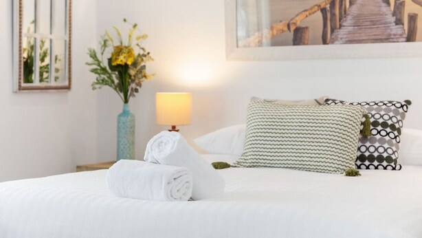 Un lit bien fait, avec du linge de lit blanc, une lampe, un vase de fleurs et un tableau représentant une jetée.