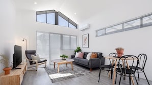 Ein modernes Wohnzimmer mit einer Couch, einem Couchtisch, Sesseln, einem Fernseher und einem großen Fenster mit Jalousien.