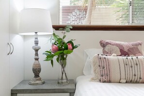 Een slaapkamer met nachtkastje, lamp, vaas met bloemen en bed met een gestreept kussen.