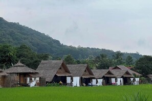 A row of traditional thatched-roof huts with a lush green field in the foreground and a forested hill in the background. - Nansilarom Boutique Resort (Pua)