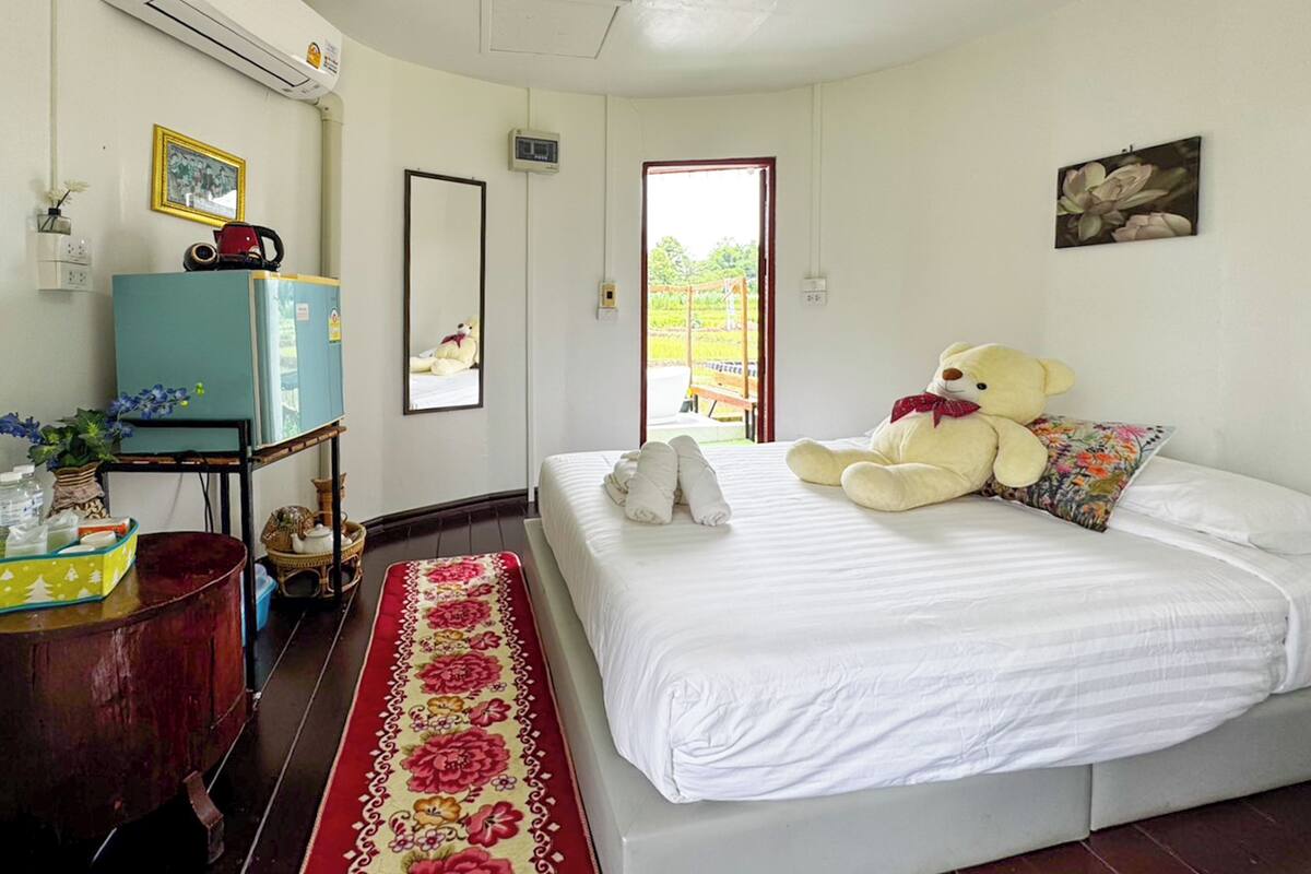 A bedroom with a bed, a large teddy bear, a mirror, a dresser, and a window with a view of greenery.