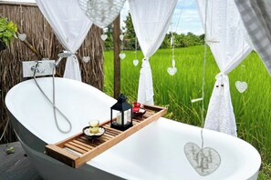 Eine Badewanne mit einem Holztablett, einem schwarzen Laternenlicht und herzförmigen Dekorationen vor einer grünen, üppigen Wiesenlandschaft.