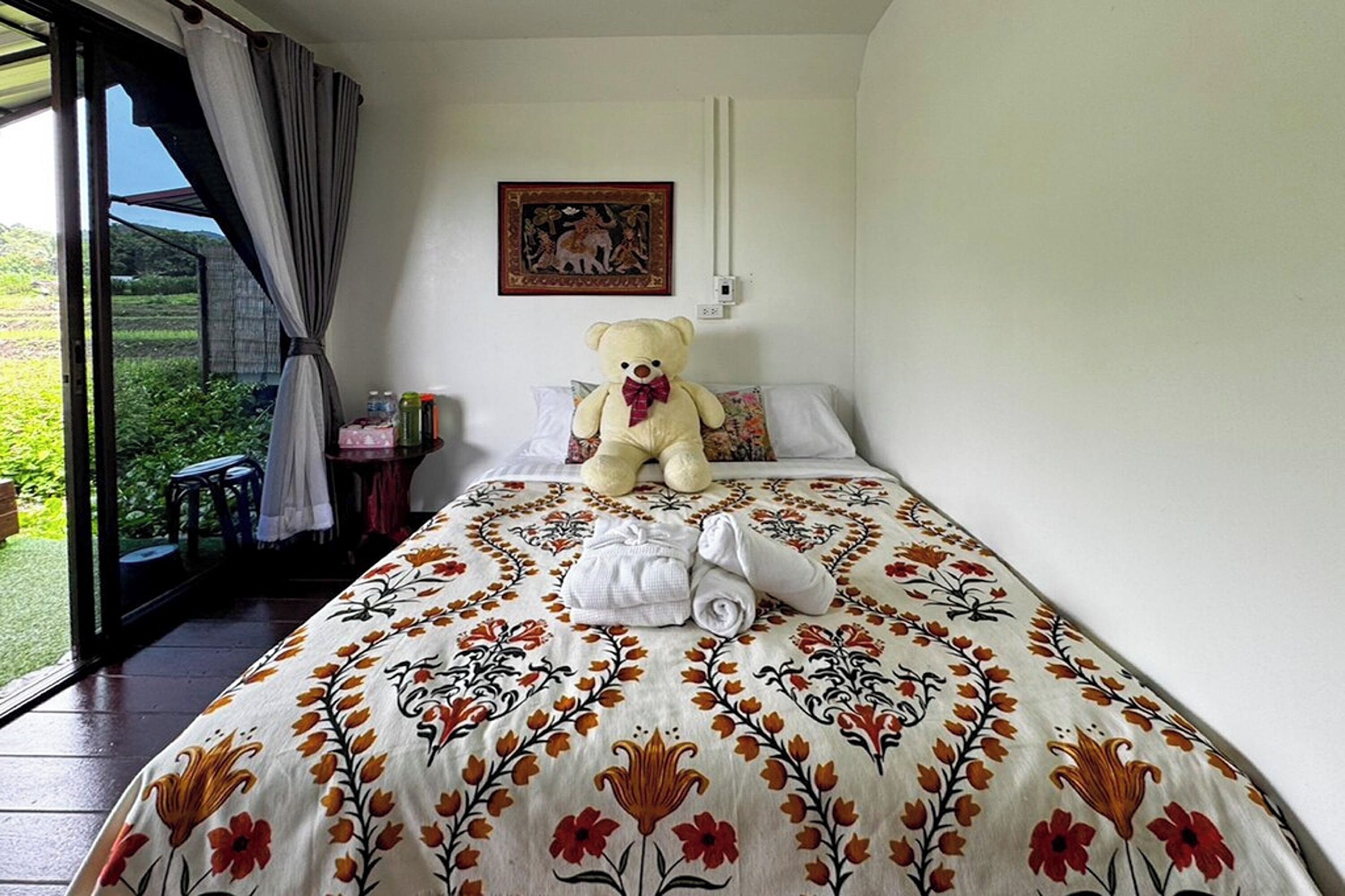 Ein Schlafzimmer mit einem großen Bett, einem Teddybären, einem Bild und Blick auf Grünfläche draußen.