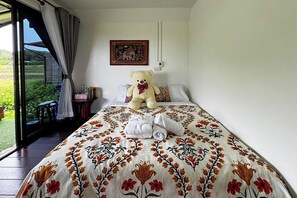 Ein Schlafzimmer mit einem großen Bett, einem Teddybären, einem Bild und Blick auf Grünfläche draußen.