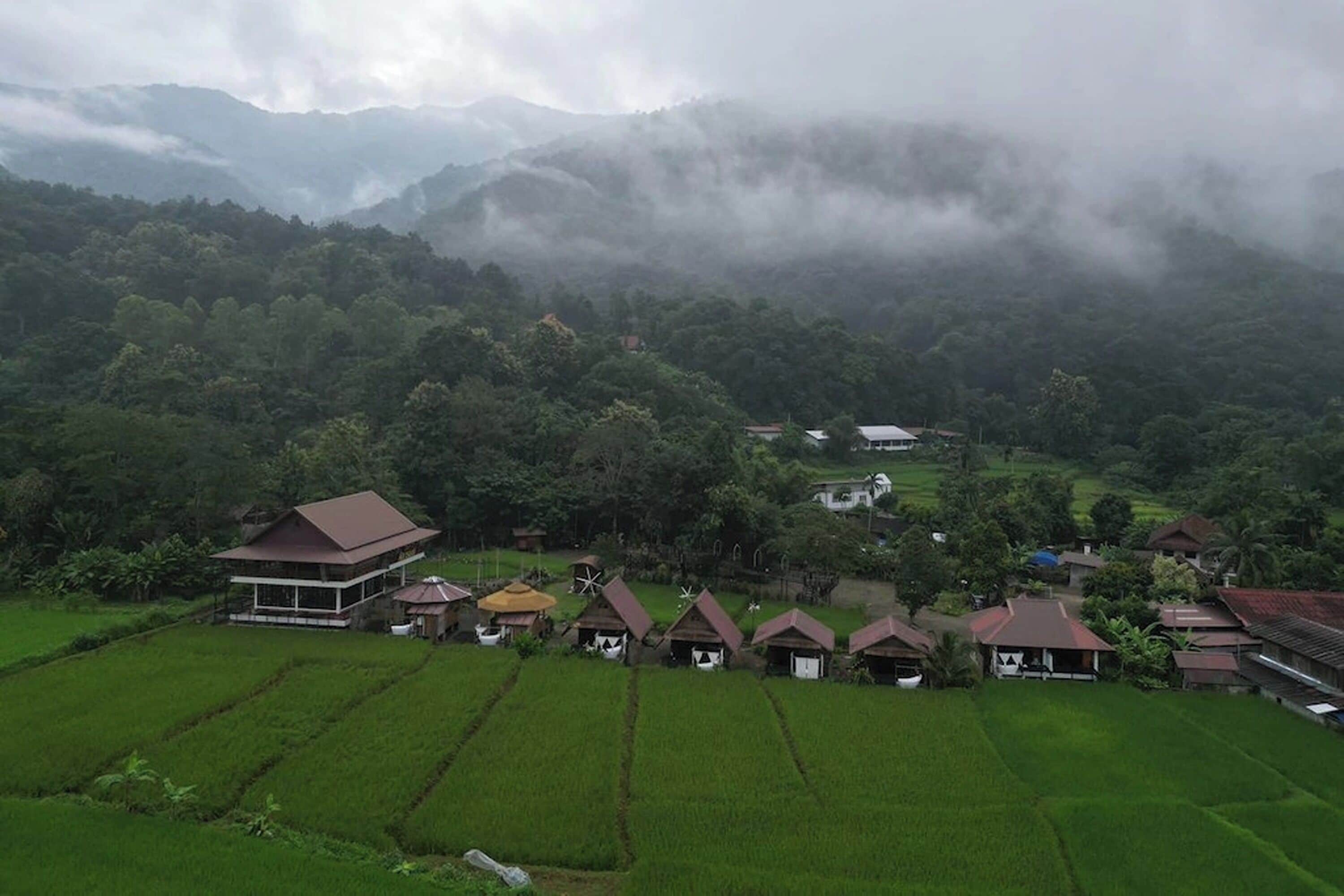Un paysage rural avec une grande maison et plusieurs bâtiments plus petits, entourés d’une végétation luxuriante et avec des montagnes en arrière-plan.